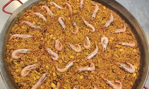 Fideua noodle paella topped with whole pink shrimp scattered across golden saffron noodles in red-handled pan at La Paella, a Spanish Restaurant in Sugar Land